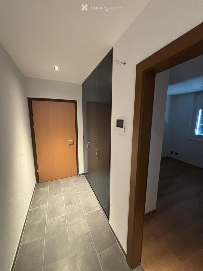 A narrow hallway with tiled flooring, white walls, and two doors: one wooden and one glass-paned. A small light fixture is mounted on the ceiling.
