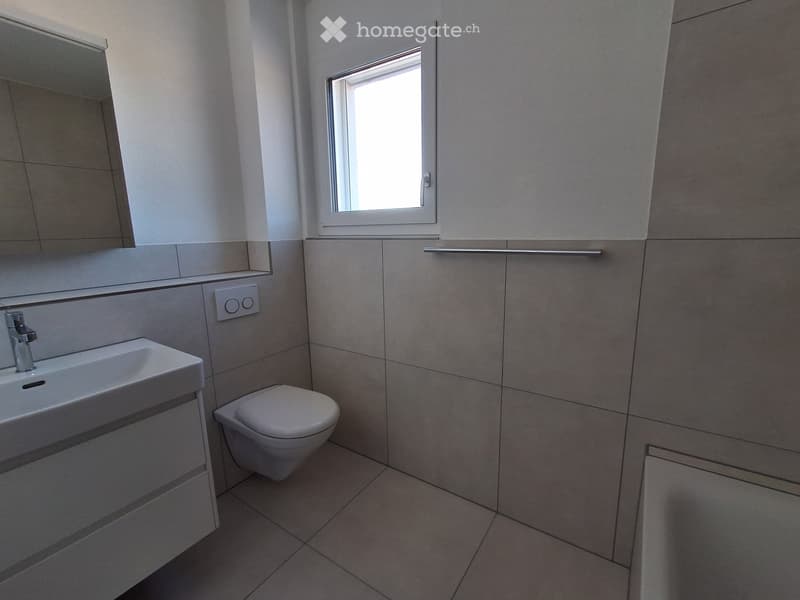 Modern bathroom with a white sink, chrome faucet, toilet, and a large mirror above the sink. Light grey tiles on walls and floor, small window providing natural light.
