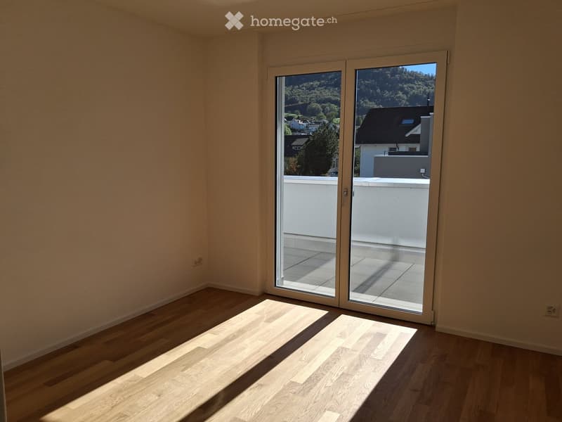 Empty room with wooden flooring and large sliding glass doors leading to a balcony, with a scenic view of trees and houses.
