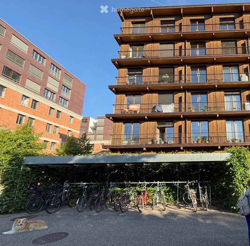 Modern multi-story apartment building with wooden balconies and large windows, surrounded by greenery and bicycles parked under a covered area.