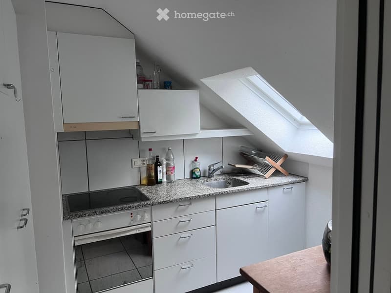 Compact kitchen with white cabinets, granite countertop, sink, oven, and various bottles on the counter.