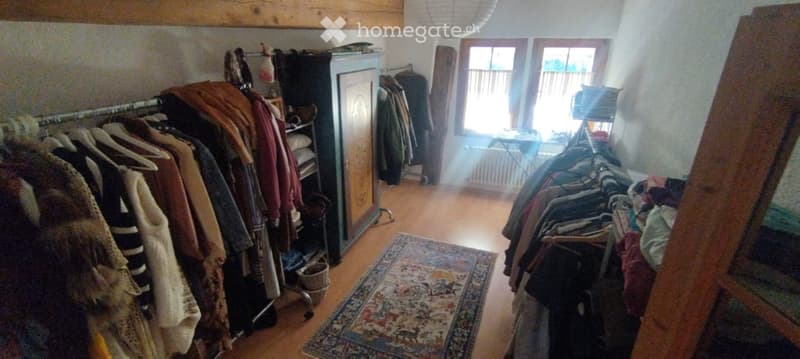 A well-lit room with wooden flooring, a large wardrobe, multiple clothing racks filled with garments, a patterned rug, and a small table near the window.