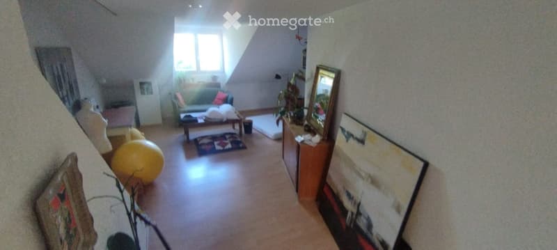 A cozy living room with a slanted ceiling, wooden floor, and large window. Features a green sofa with colorful cushions, a wooden coffee table, a large yellow exercise ball, a mirror, and several picture frames on the walls.