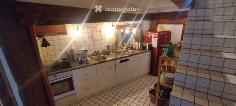 A cozy kitchen with white tiled walls and floor, wooden cabinets, a red fridge, and various kitchen appliances and utensils on the counter and shelves.