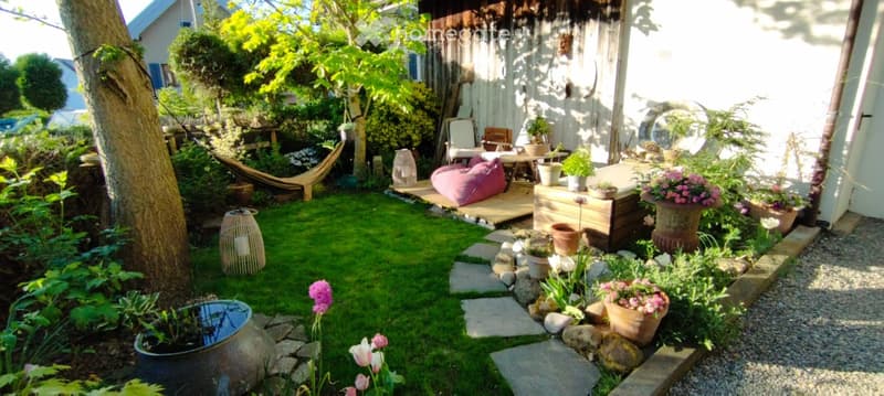 A small, well-maintained garden with a grassy area, stone pathway, potted plants, and a wooden deck with seating and a hammock.