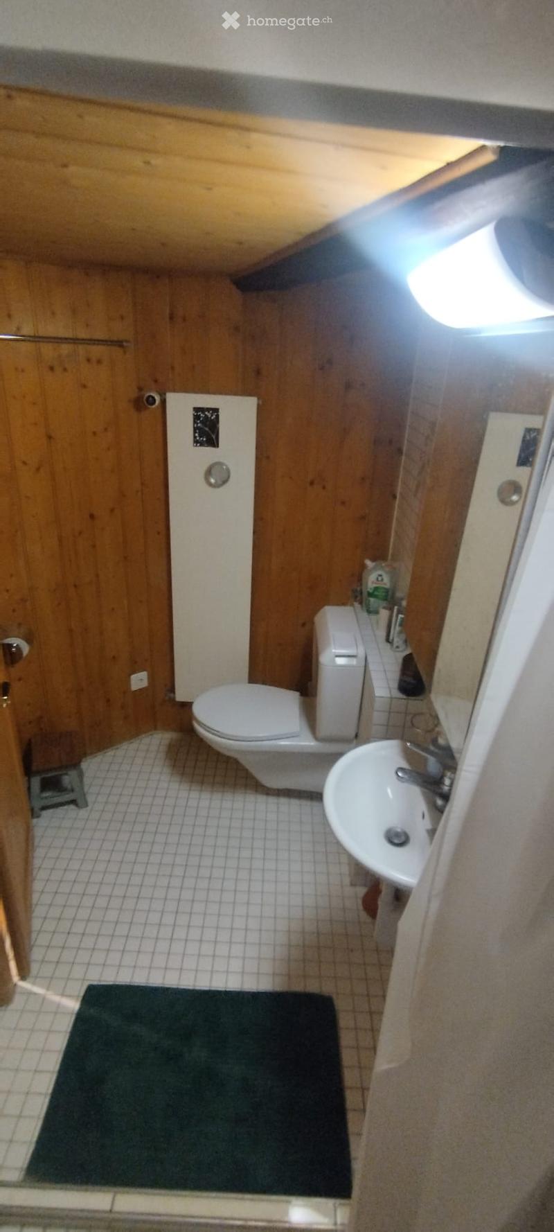 Compact bathroom with wooden walls, white tiled floor, toilet, sink, and small stool near the door.