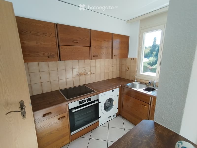 Compact kitchen with wooden cabinets, tiled backsplash, stainless steel sink, oven, washing machine, and a small wooden table.