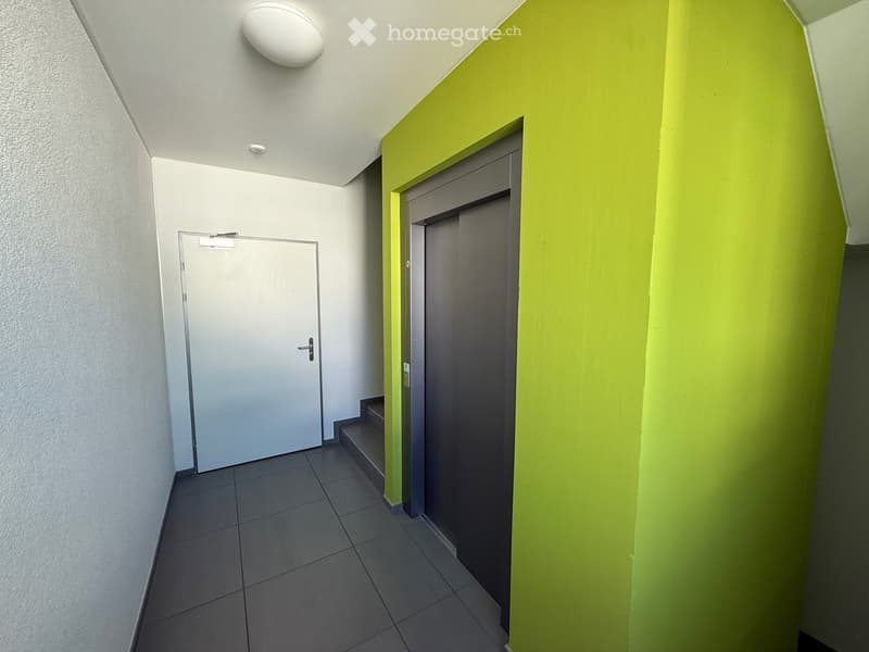 A modern hallway with tiled flooring, white walls, and a bright green accent wall. Features include a closed door on the left, an open doorway leading to a staircase on the right, and a ceiling light fixture.