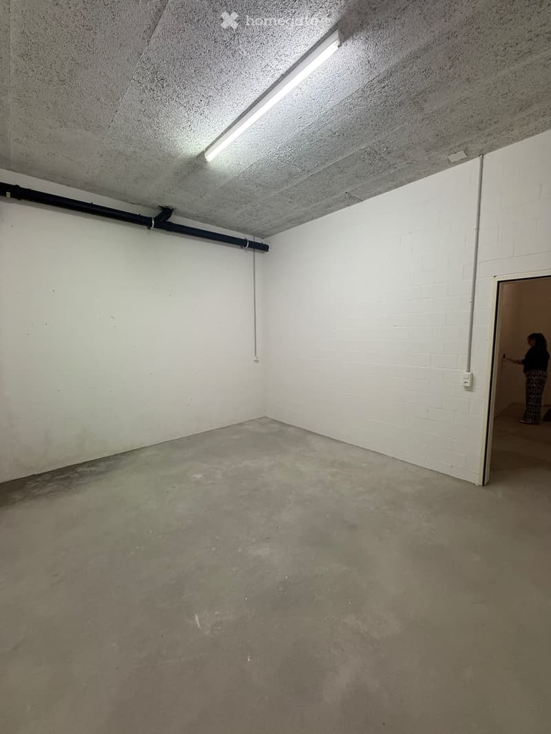 Empty room with white brick walls, concrete floor, and exposed ceiling with fluorescent lighting. A doorway leads to another area where a person is visible.