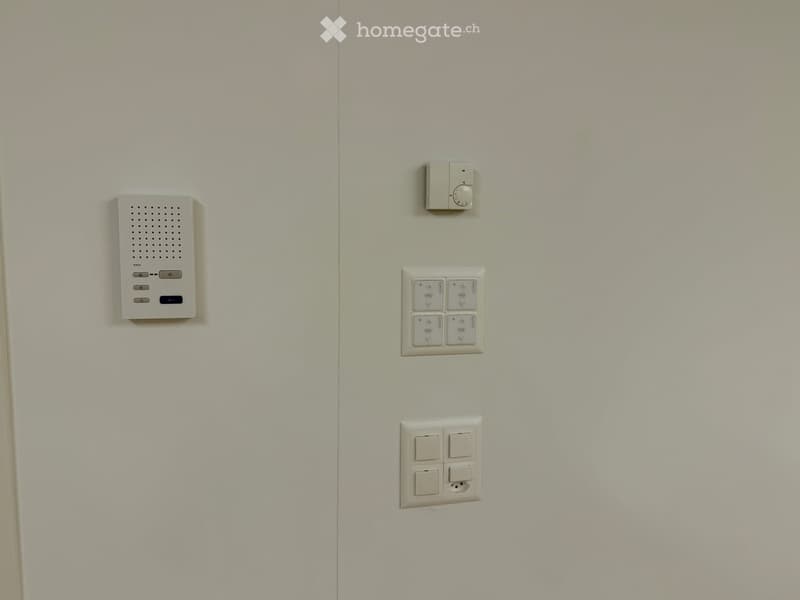 A modern hallway with a white wall featuring several electronic devices including a speaker, a light switch, and a power outlet.
