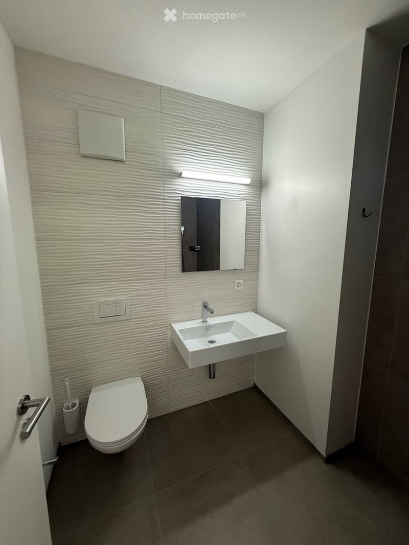 Modern bathroom with a white toilet, a white sink with a chrome faucet, a large mirror above the sink, and a light fixture integrated into the wall. The walls feature a textured, light-colored tile design, and the floor is covered with dark tiles.