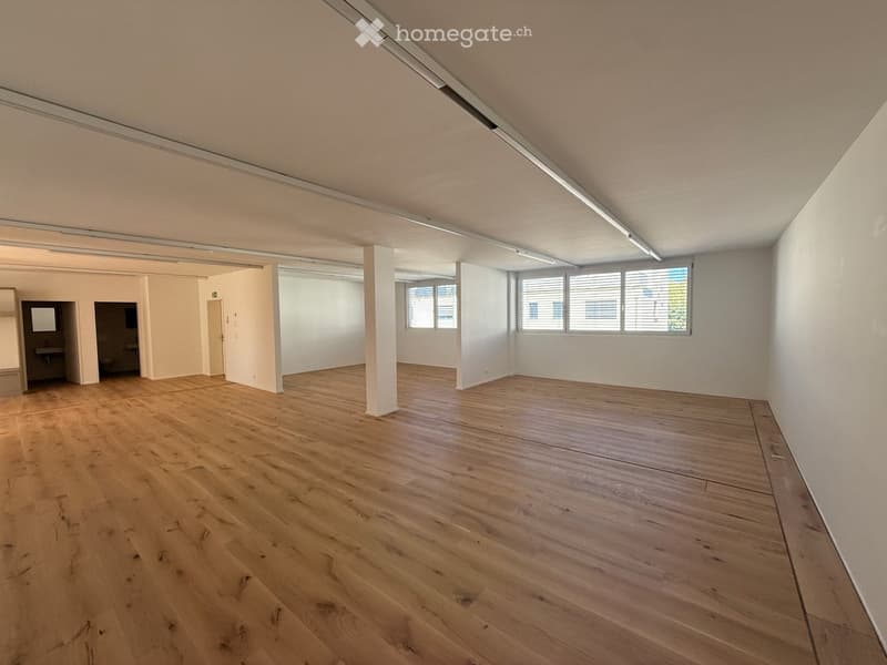 Empty room with wooden flooring, white walls, and large windows with blinds. The space includes a small adjacent area with a sink and mirror.