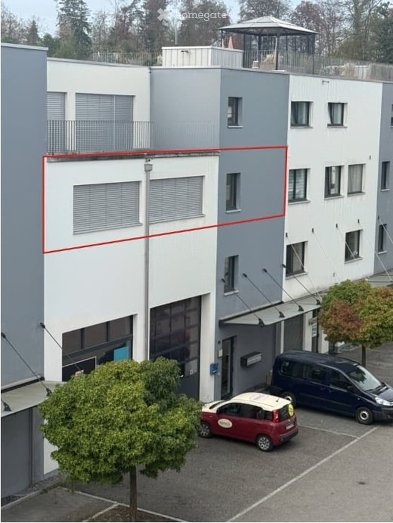 Modern multi-story building with white and grey facade, multiple windows, and a small balcony on the top floor. Two cars are parked in front of the building.