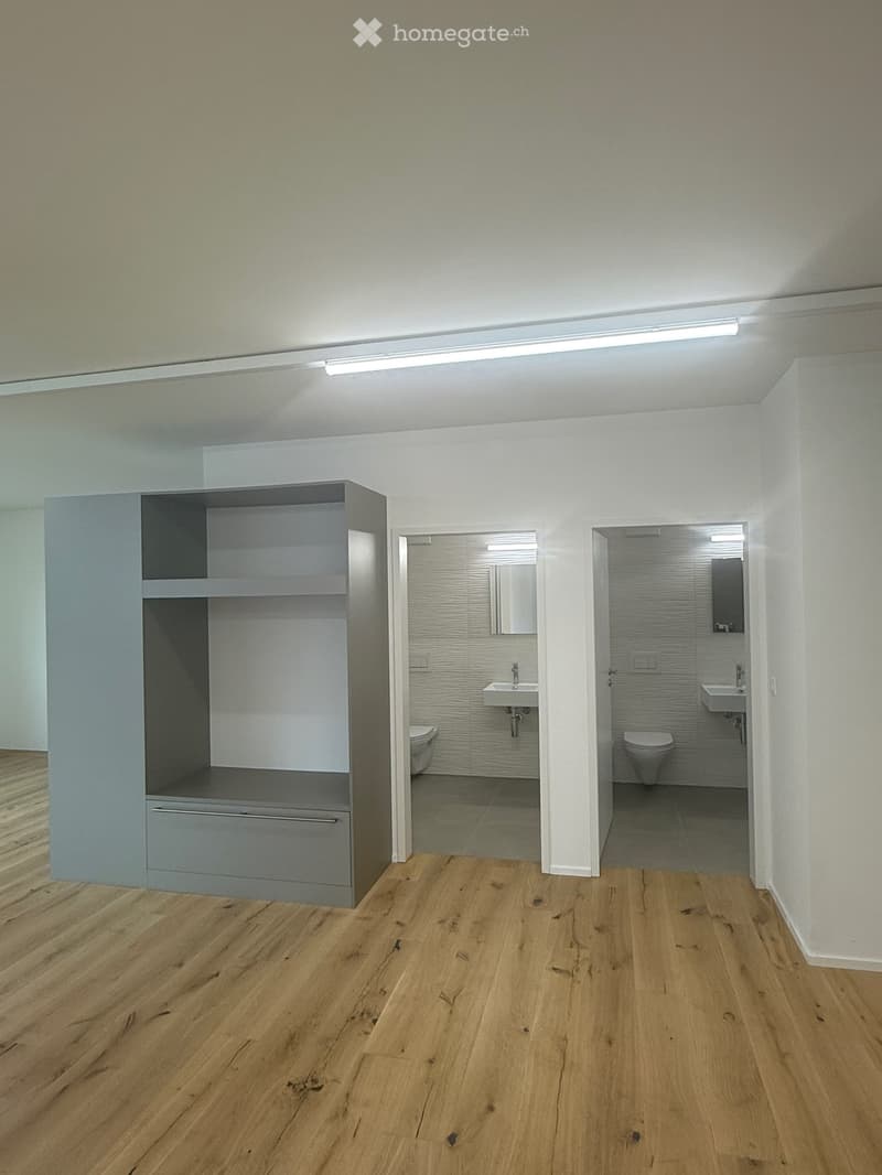 Empty room with wooden flooring, white walls, and built-in storage unit. Two bathrooms are visible through open doorways, each containing a sink, toilet, and mirror.