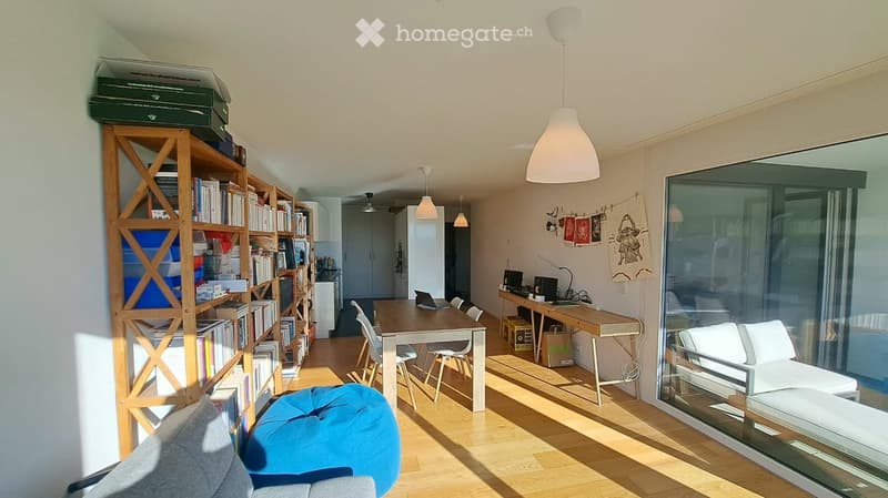 Bright, open-plan living and dining area with wooden flooring, large windows, a sofa, a blue ball chair, a dining table with chairs, a desk with a laptop, and a bookshelf filled with books.