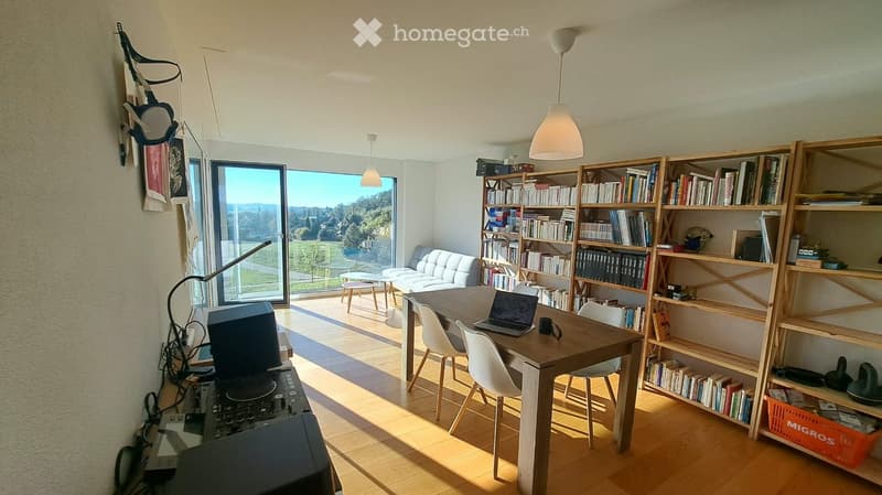 Bright, open living space with wooden flooring, large windows offering a scenic view, a sofa, a dining table with chairs, a laptop, and a bookshelf filled with books.