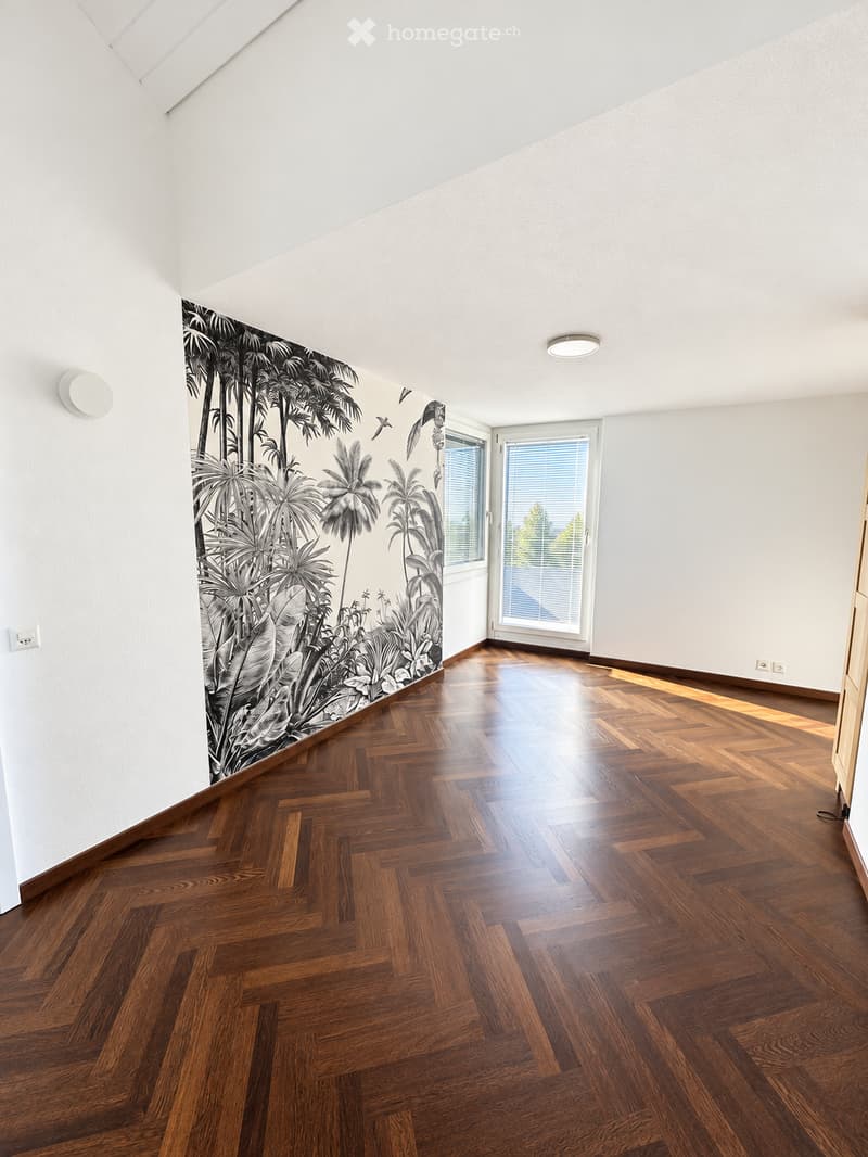 Empty room with wooden herringbone flooring, white walls, large window with blinds, and a mural of tropical plants on one wall.