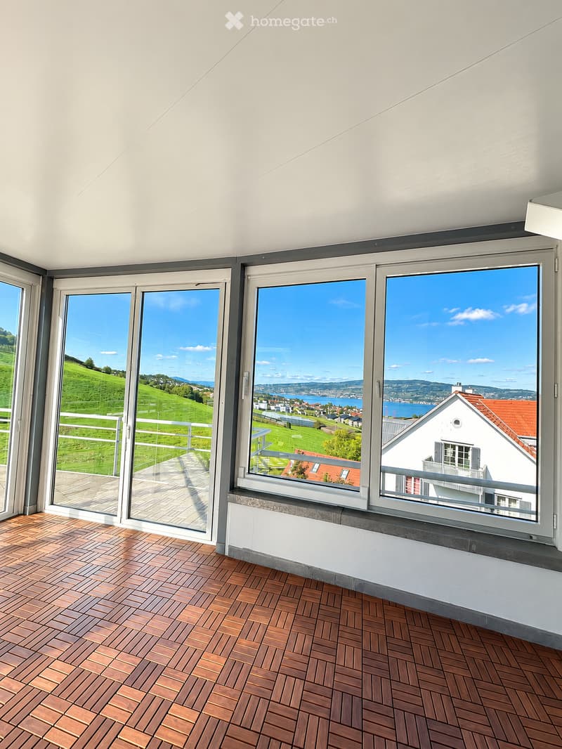 Bright balcony with large windows offering scenic views of green fields and a body of water. Wooden decking and white railings.
