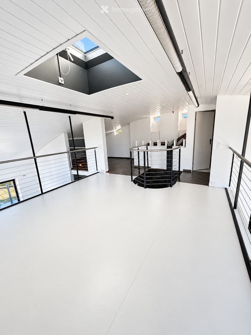 Modern, minimalist hallway with white walls, white flooring, and a sleek black metal staircase with a glass railing. The ceiling features a skylight and recessed lighting.