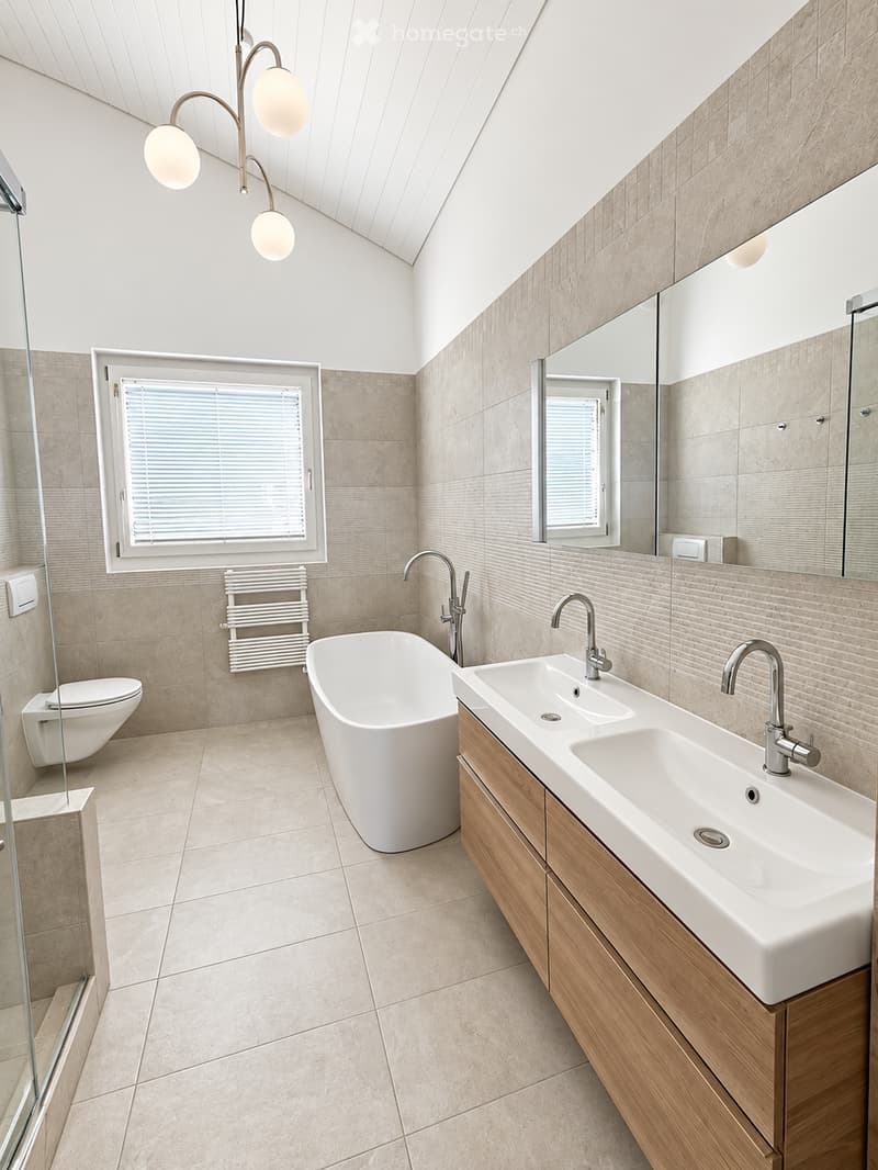 Modern bathroom with a freestanding bathtub, double sink, toilet, and large mirror. Features include a radiator, modern lighting fixture, and large windows with blinds.