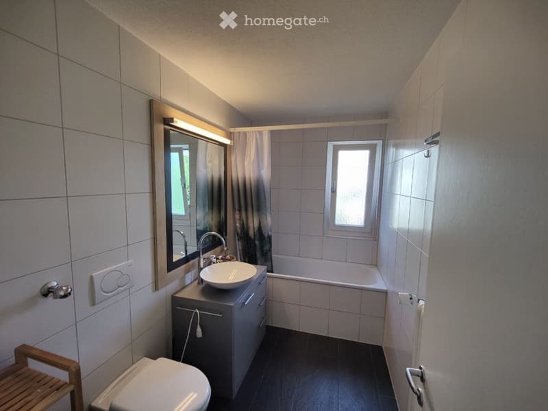 Modern bathroom with a white sink, bathtub, toilet, and large mirror. The room features tiled walls and a window providing natural light.