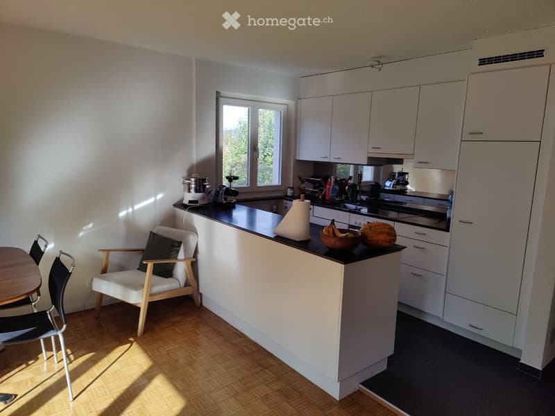 Modern kitchen with white cabinets, black countertops, and a small dining area featuring a chair and table. The kitchen includes various appliances and a bowl of fruit on the counter.