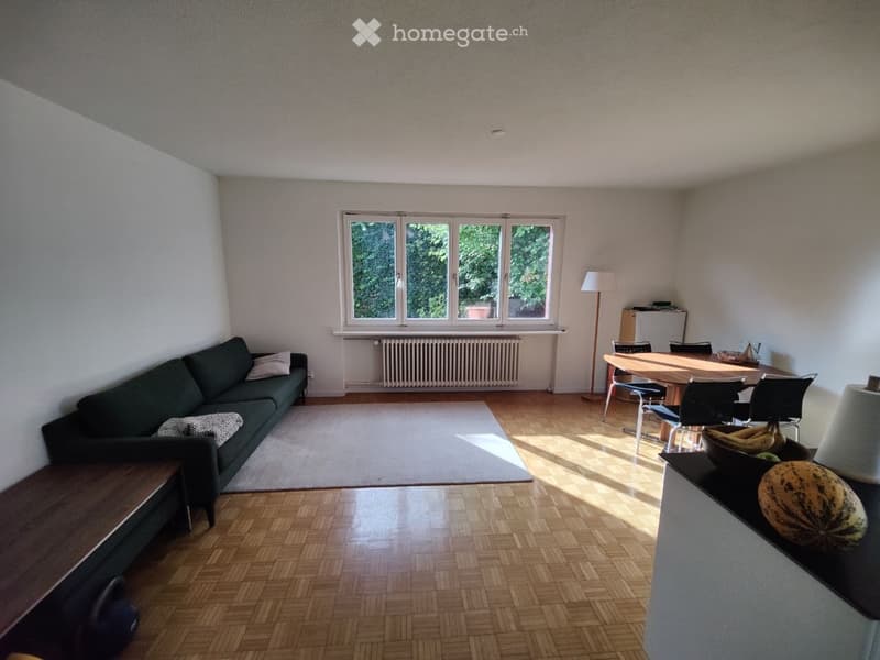 A well-lit living and dining area with wooden flooring, a dark green sofa, a dining table with chairs, and a floor lamp. The room features large windows with a view of greenery outside.