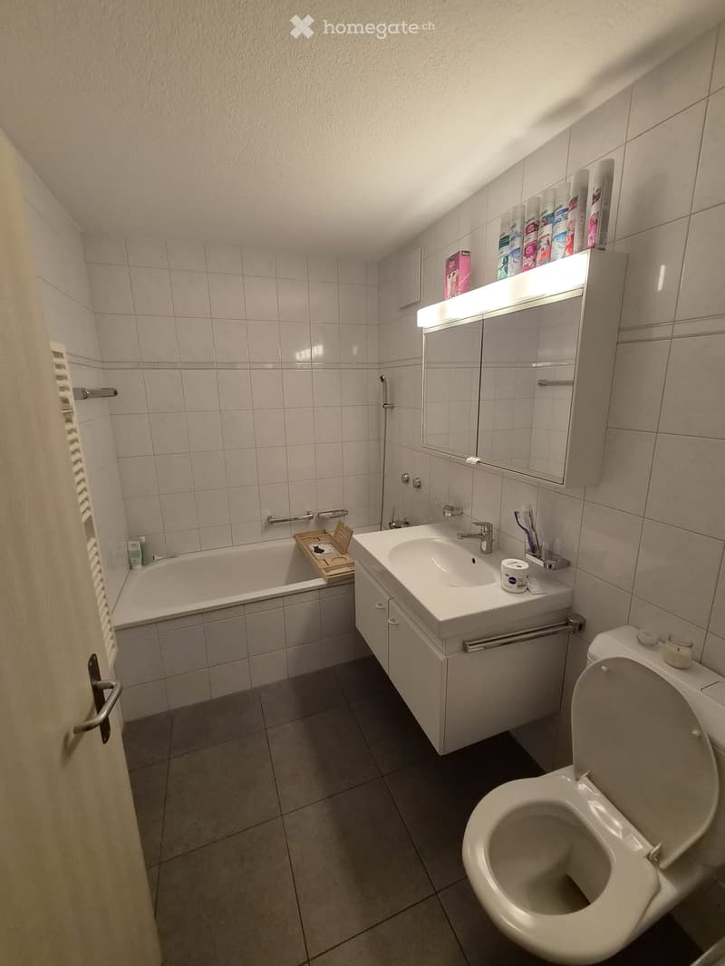 Modern bathroom with white tiles, a bathtub, sink, toilet, mirror, and a built-in cabinet with a light fixture above the mirror. The sink has a faucet and toothbrush holder, and the bathtub has a wooden tray with items on it.