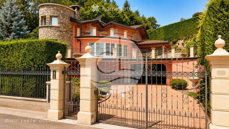 A large, elegant house with a stone tower, pink facade, and multiple balconies surrounded by lush greenery and a decorative iron gate.