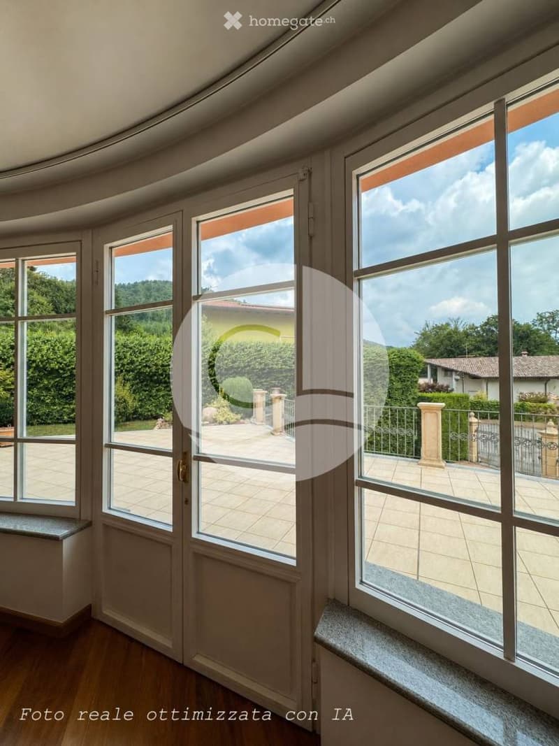 Bright balcony with large windows offering a scenic view of a garden and surrounding landscape. Wooden flooring and tiled balcony area visible.