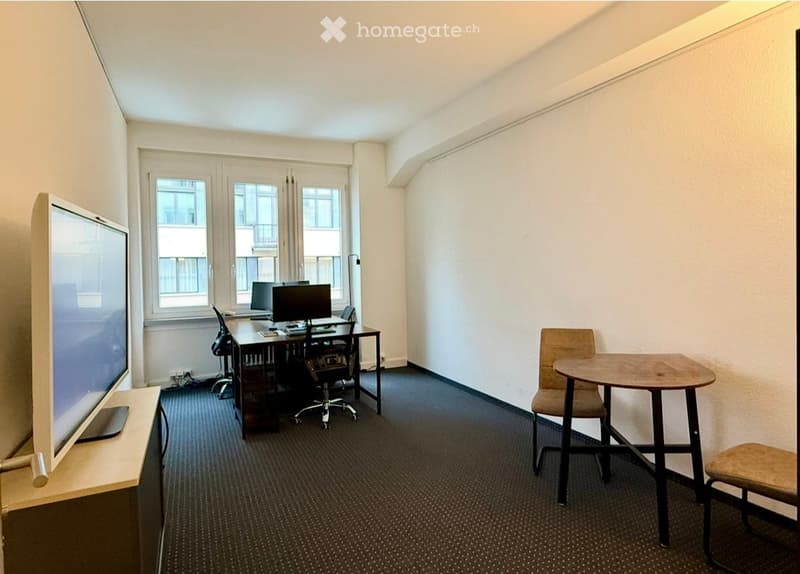 Office space with a large screen, a desk with a computer, chairs, and a small round table with a stool.