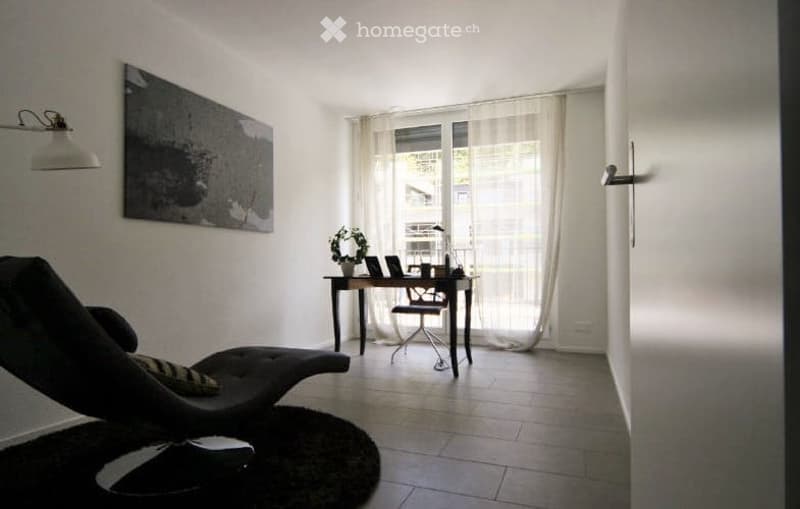 A modern, minimalist room with a black lounge chair, a small desk with a laptop, a lamp, and a potted plant. The room features large windows with sheer curtains and a sleek, contemporary design.