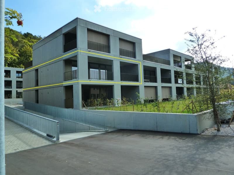 Modern multi-story building with grey concrete facade, multiple balconies, and a raised ground floor entrance.