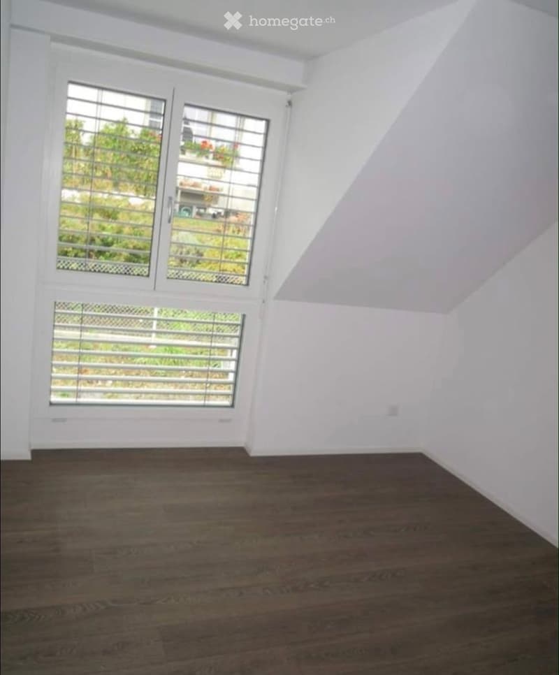 Empty room with wooden flooring and white walls, featuring large windows with blinds and a view of greenery outside.