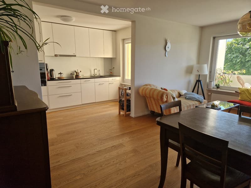 A modern, open-plan living and dining area with a striped sofa, wooden dining table, and kitchen area featuring white cabinets and stainless steel appliances.