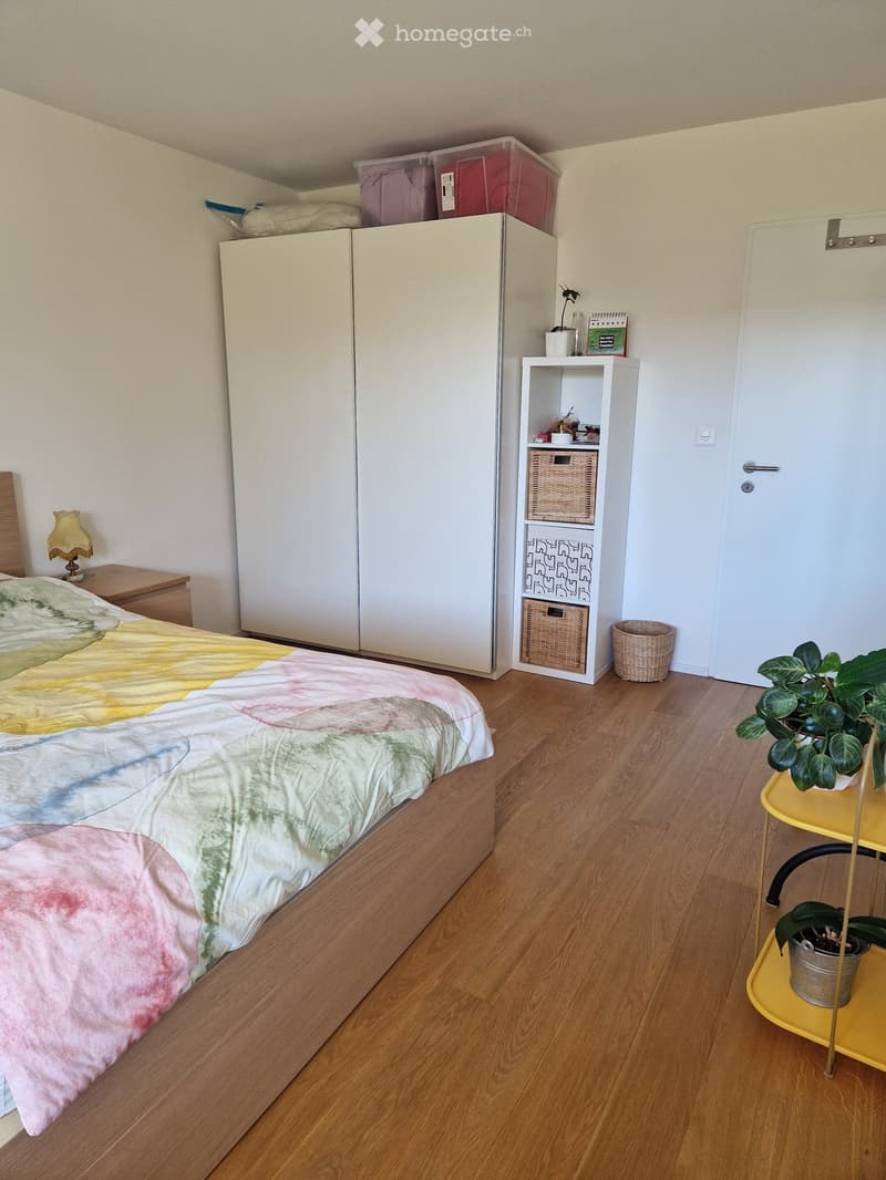 A tidy bedroom with a wooden bed, colorful tie-dye duvet, nightstand with lamp, large white wardrobe, small shelf with baskets, and a yellow side table with potted plants.