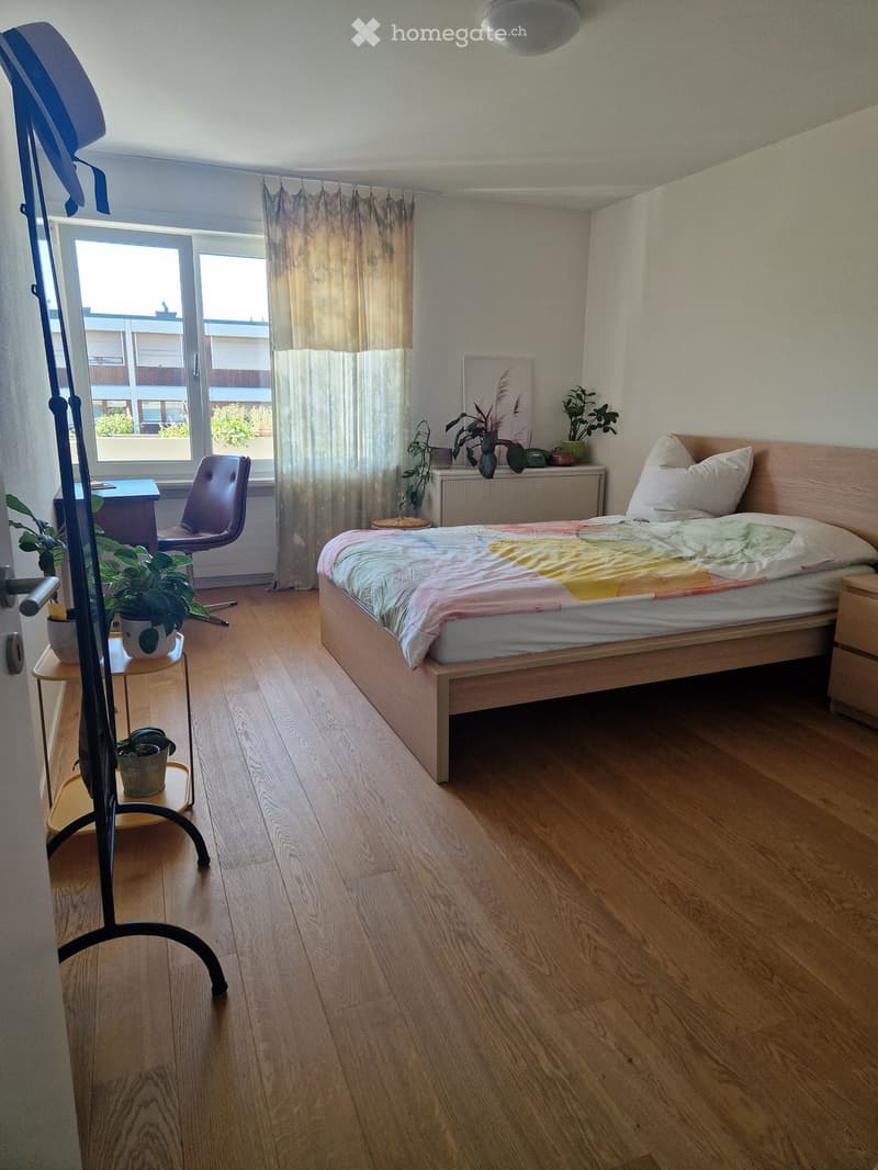Cozy bedroom with wooden flooring, a bed with colorful bedding, a small table with plants, a chair, and a radiator near the window.