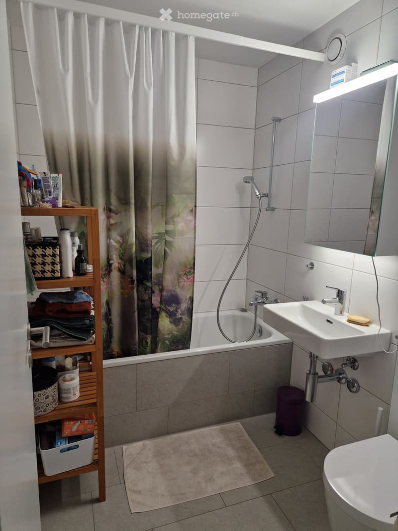 Modern bathroom with a bathtub, sink, toilet, and a wooden shelf holding towels and toiletries. Features a shower curtain, a mirror with built-in lighting, and a small trash bin.