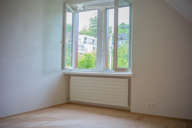 Empty room with wooden flooring, white walls, large windows, radiator, and electrical outlets.