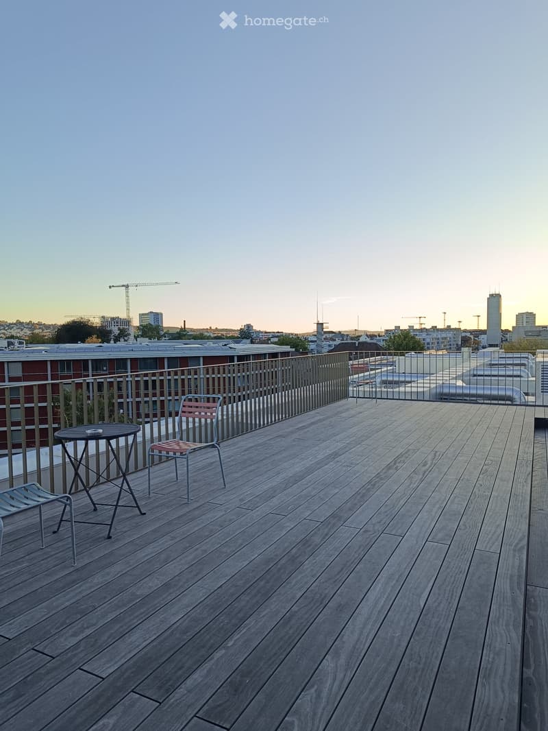 Wooden deck with a small table and two chairs, metal railing, cityscape view in the background.