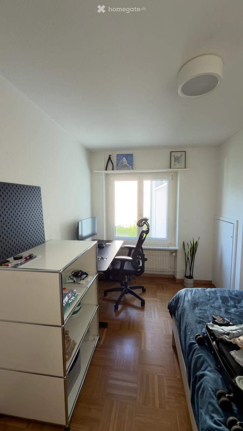 A modern bedroom with a bed, desk, chair, and a cabinet. The room features wooden flooring and large windows letting in natural light.