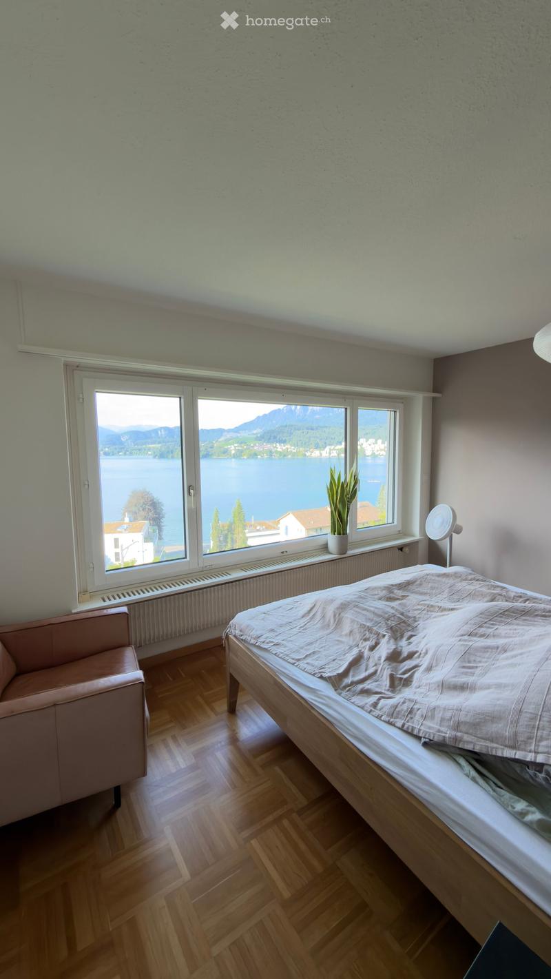 Bright bedroom with wooden flooring, large window offering a scenic lake and mountain view, bed, sofa, fan, and potted plant on the windowsill.