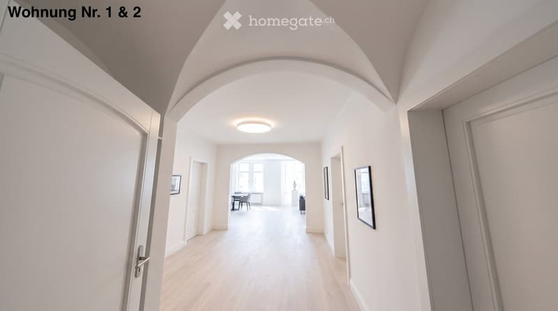 Bright hallway with wooden flooring, white walls, and recessed lighting. Features arched doorways leading to adjacent rooms, with visible furniture in the background.