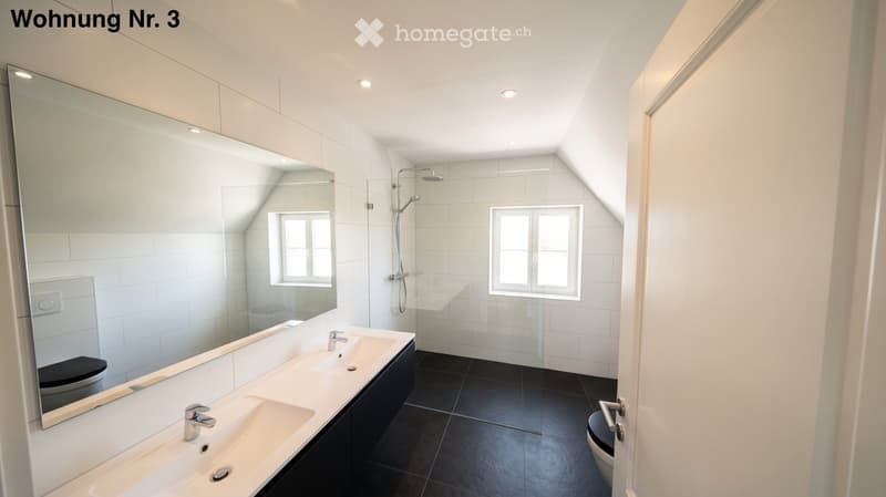 Modern bathroom with two sinks, a large mirror, a toilet, and a shower area with a window. The space features white tiles and black flooring.