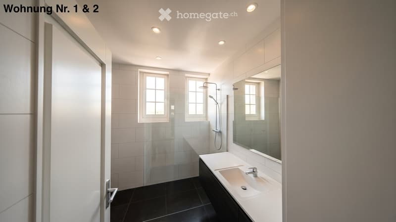 Modern bathroom with white tiles, black floor, large mirror, sink, and glass-enclosed shower area.