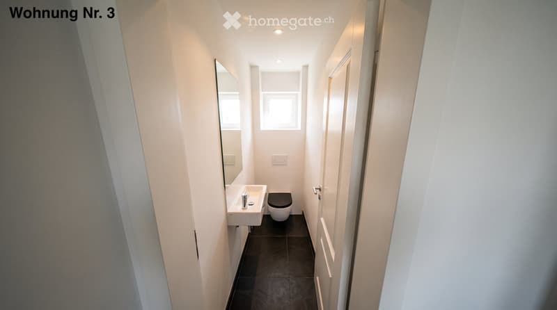 Modern, compact bathroom with white walls, dark tiled floor, a sink, toilet, and a mirror.