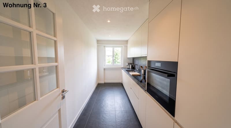 Modern kitchen with white cabinets, dark countertops, and stainless steel appliances, including a sink, faucet, and oven. The space features large windows and a sleek design.