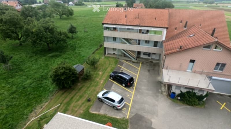 Aerial view of a two-story residential building with a parking area, surrounded by green fields and trees.