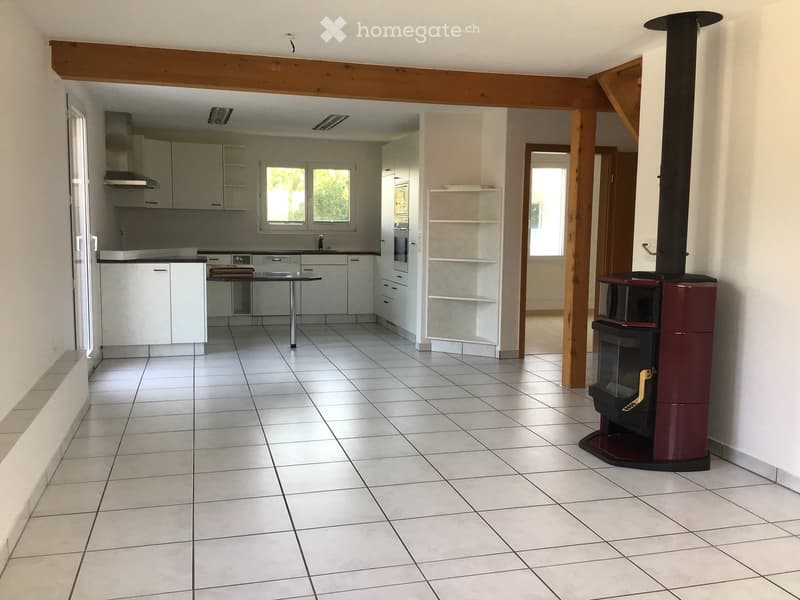 Open-plan kitchen and living area with white tiled flooring, white kitchen cabinets, a central table, and a wood stove.