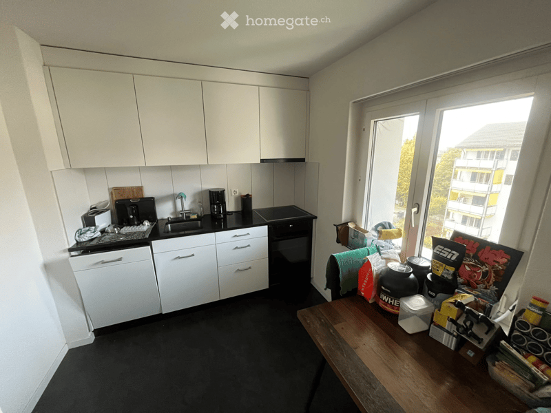 Modern kitchen with white cabinets, black countertops, and a large window overlooking a building. Includes a sink, faucet, oven, coffee maker, toaster, and a wooden table with various items on it.